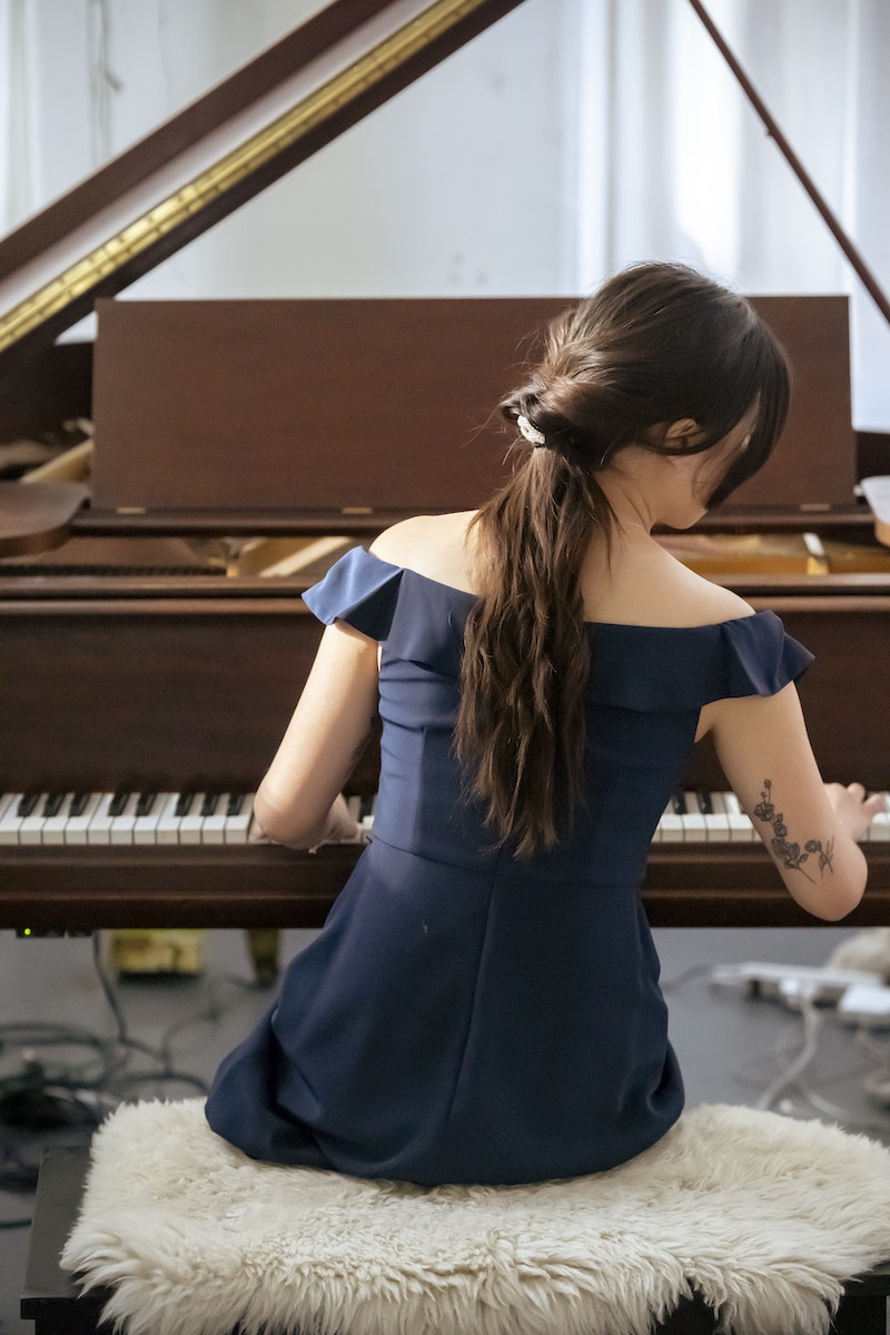 Back view of unrecognizable lady in elegant dress playing piano while sitting on pouf in light room